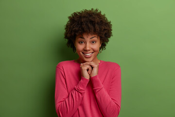 Wall Mural - Cute young dark skinned curly haired female model keeps hands pressed together under chin, smiles gently at camera, shows white perfect teeth, wears rosy clothes, models against green background