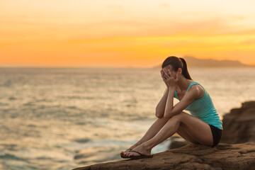 Sad crying young woman with hands on face sitting alone at the beach during a orange sunset. Depression and heartbreak concept.