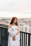 Fototapeta Krajobraz - Beautiful girl on the terrace in a white blanket at sunset. Girl posing background fashion. Mood girl wrapping with blanket on balcony. Terrace mood.