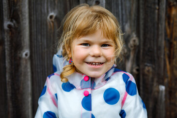 Outdoor portrait of happy smiling little toddler girl wearing rain jacket on rainy cloudy day. Cute healthy child in colorful clothes outdoor activity