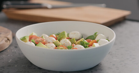 add spices into salad with kale, avocado, mozzarella and tomatoes in white bowl on kitchen countertop