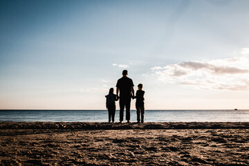 father holding hands with son and daughter looking at the water while standing on the beach