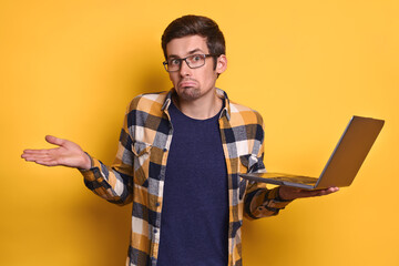 Portrait of confused frustrated man in glasses and casual clothes holding laptop computer on yellow studio wall, making don't know gesture, doesn't understand. Human emotion and technology concept