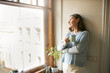 © shurkin_son - Portrait of thoughtful retired mature woman in casual clothes standing by large window, leaning on white wall, looking outside, drinking tea. People, social distancing and quarantine concept