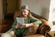 People, aging and electronic gadgets. Modern attractive middle aged woman with long gray hair relaxing at home using digital tablet to communicate online with family, looking at screen with smile