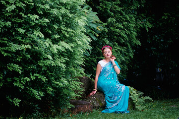 Wall Mural - Beautiful young caucasian woman in traditional indian clothing with bridal makeup and jewelry. Bollywood dancer in Sari and henna on hands walking on green garden.