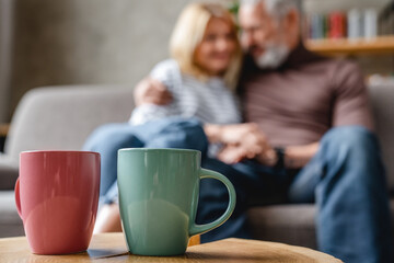 Wall Mural - Two coffee cups on table with senior couple relaxing on background on couch