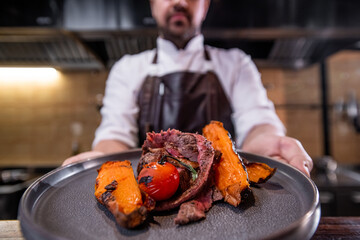 Close-up of chef showing roasted meat with sweet potatoes and tomatoes on plate