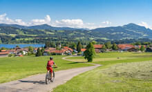 Pretty Senior Woman Riding Her Electrical Mountain Bike In Warm  Sunlight And Enjoying The Spectacular View Over The Allgau Alps Near Moosdorf With Rottach Lake And Gruenten Summit, Bavaria, Germany