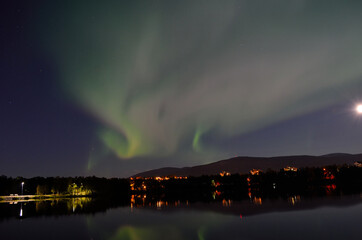 majestic aurora borealis, northern light over calm mirror lake at night with buildings in the backgr