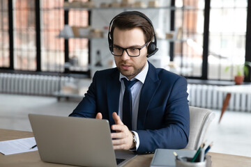 Canvas Print - Concentrated Caucasian businessman in wireless headset talk on video call on webcam on laptop in office, serious male employer in earphones speak on web conference using modern computer at workplace