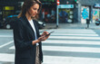 © A_B_C - Woman using cellphone and crossing street, young hipster girl in business wear holding smartphone gadget using for online booking taxi cab waiting for transport near city road with traffic