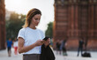 © A_B_C - Portrait of beautiful traveler woman reading message on smart phone walking arc de Triomphe of Barcelona, tourist girl using mobile phone in sun summer day park outdoors