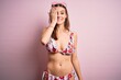 © Krakenimages.com - Young beautiful woman on vacation wearing bikini and sunglasses over pink background covering one eye with hand, confident smile on face and surprise emotion.