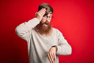 Wall Mural - Handsome Irish redhead man with beard wearing casual sweater and glasses over red background Looking at the watch time worried, afraid of getting late