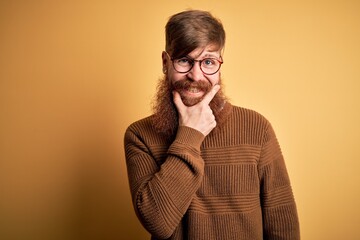 Poster - Handsome Irish redhead man with beard wearing glasses and winter sweater over yellow background looking confident at the camera smiling with crossed arms and hand raised on chin. Thinking positive.