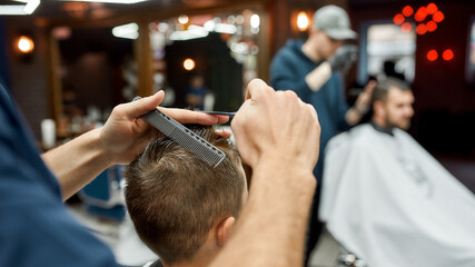 Cropped photo of barber with scissors and hair comb in hands making a haircut for little boy in the modern barbershop. Selective focus