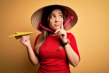 Canvas Print - Tourist woman wearing traditional asian rice paddy straw hat holding paper plane for a trip serious face thinking about question, very confused idea