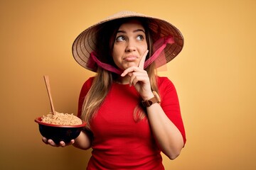 Poster - Young woman wearing asian hat and eating chinese noodles over yellow isolated background serious face thinking about question, very confused idea