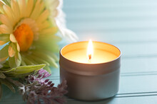 Lighted And Scented Candle In A Tin Holder With Flowers On The Side, All On Teal Boards Table Background With Back Window Light Area As Copy Space.  Horizontal Photo With A Front View