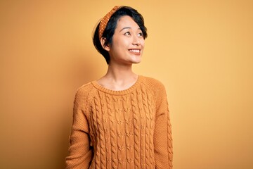 Poster - Young beautiful asian girl wearing casual sweater and diadem standing over yellow background looking away to side with smile on face, natural expression. Laughing confident.