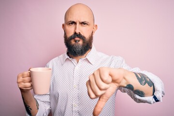 Sticker - Handsome bald man with beard and tattoo drinking cup of coffee over pink background with angry face, negative sign showing dislike with thumbs down, rejection concept