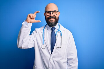 Wall Mural - Handsome bald doctor man with beard wearing glasses and stethoscope over blue background smiling and confident gesturing with hand doing small size sign with fingers looking and the camera. Measure