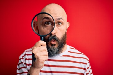 Canvas Print - Handsome detective bald man with beard using magnifying glass over red background scared in shock with a surprise face, afraid and excited with fear expression