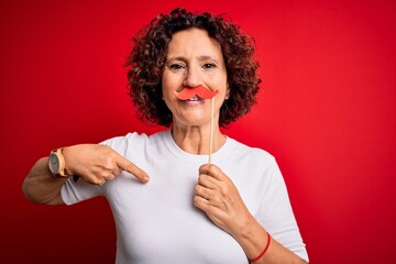 Poster - Middle age curly hair woman holding funny mustache over isolated red background with surprise face pointing finger to himself