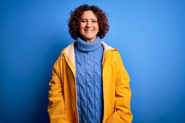 Poster - Middle age curly hair woman wearing rain coat standing over isolated blue background with a happy and cool smile on face. Lucky person.