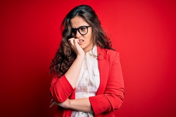 Poster - Young beautiful woman with curly hair wearing jacket and glasses over red background looking stressed and nervous with hands on mouth biting nails. Anxiety problem.