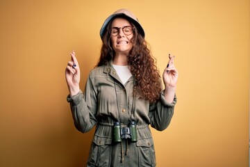 Poster - Young beautiful tourist woman on vacation wearing explorer hat and binoculars gesturing finger crossed smiling with hope and eyes closed. Luck and superstitious concept.