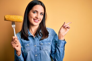 Young woman with blue eyes holding fork with fresh cob corn standing over yellow background very happy pointing with hand and finger to the side