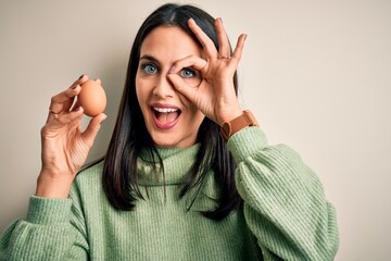 Wall Mural - Young brunette woman with blue eyes holding fresh raw egg over isolated background with happy face smiling doing ok sign with hand on eye looking through fingers