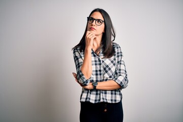 Wall Mural - Young brunette woman with blue eyes wearing casual shirt and glasses over white background with hand on chin thinking about question, pensive expression. Smiling with thoughtful face. Doubt concept.