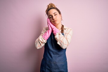 Young beautiful blonde cleaner woman doing housework wearing arpon and gloves sleeping tired dreaming and posing with hands together while smiling with closed eyes.