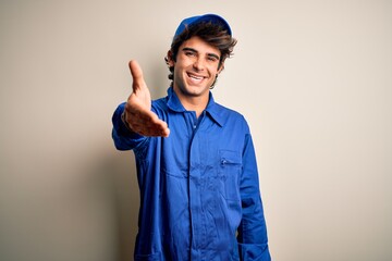 Wall Mural - Young mechanic man wearing blue cap and uniform standing over isolated white background smiling friendly offering handshake as greeting and welcoming. Successful business.