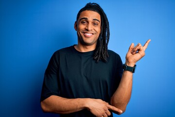 Wall Mural - Young handsome african american afro man with dreadlocks wearing casual t-shirt with a big smile on face, pointing with hand and finger to the side looking at the camera.