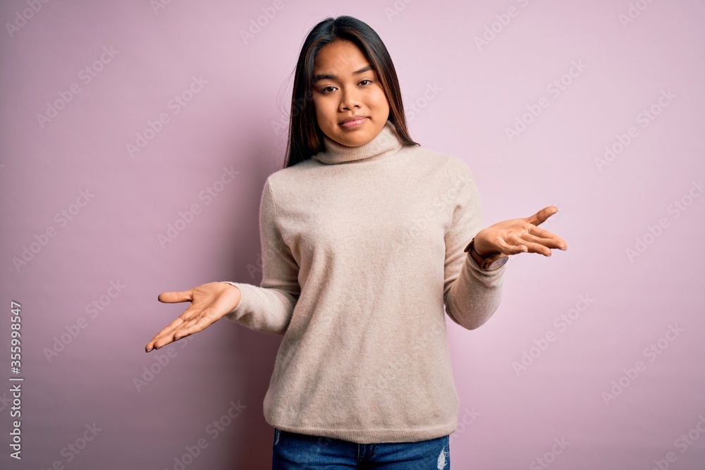 Young beautiful asian girl wearing casual turtleneck sweater over isolated pink background clueless and confused expression with arms and hands raised. Doubt concept.