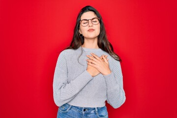 Canvas Print - Young beautiful brunette woman wearing casual sweater over red background smiling with hands on chest with closed eyes and grateful gesture on face. Health concept.