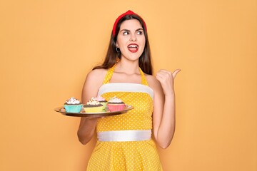 Canvas Print - Young beautiful pin up woman wearing 50s fashion vintage dress holding tray with cupcakes pointing and showing with thumb up to the side with happy face smiling