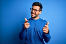 Young Handsome Man With Beard Wearing Casual Sweater And Glasses Over Blue Background Pointing Fingers To Camera With Happy And Funny Face. Good Energy And Vibes.