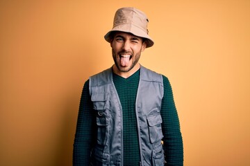 Canvas Print - Handsome tourist man with beard on vacation wearing explorer hat over yellow background sticking tongue out happy with funny expression. Emotion concept.