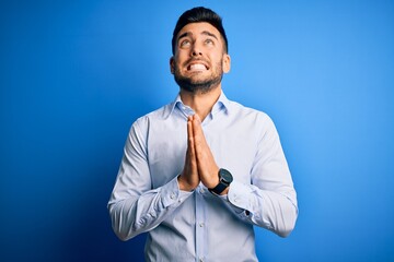 Wall Mural - Young handsome man wearing elegant shirt standing over isolated blue background begging and praying with hands together with hope expression on face very emotional and worried. Begging.