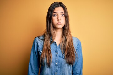 Poster - Young beautiful girl wearing casual denim shirt standing over isolated yellow background puffing cheeks with funny face. Mouth inflated with air, crazy expression.