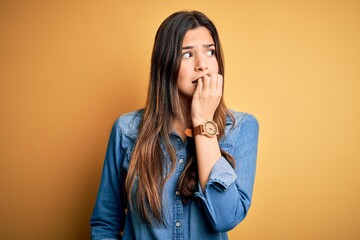 Poster - Young beautiful girl wearing casual denim shirt standing over isolated yellow background looking stressed and nervous with hands on mouth biting nails. Anxiety problem.