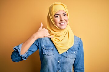 Wall Mural - Young beautiful girl wearing muslim hijab standing over isolated yellow background smiling doing phone gesture with hand and fingers like talking on the telephone. Communicating concepts.