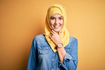 Poster - Young beautiful girl wearing muslim hijab standing over isolated yellow background looking confident at the camera smiling with crossed arms and hand raised on chin. Thinking positive.