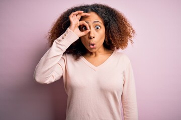 Wall Mural - Young african american woman with afro hair wearing casual sweater over pink background doing ok gesture shocked with surprised face, eye looking through fingers. Unbelieving expression.