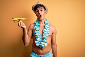 Poster - Young tourist man on vacation wearing swimwear and hawaiian lei flowers holding paper plane scared in shock with a surprise face, afraid and excited with fear expression